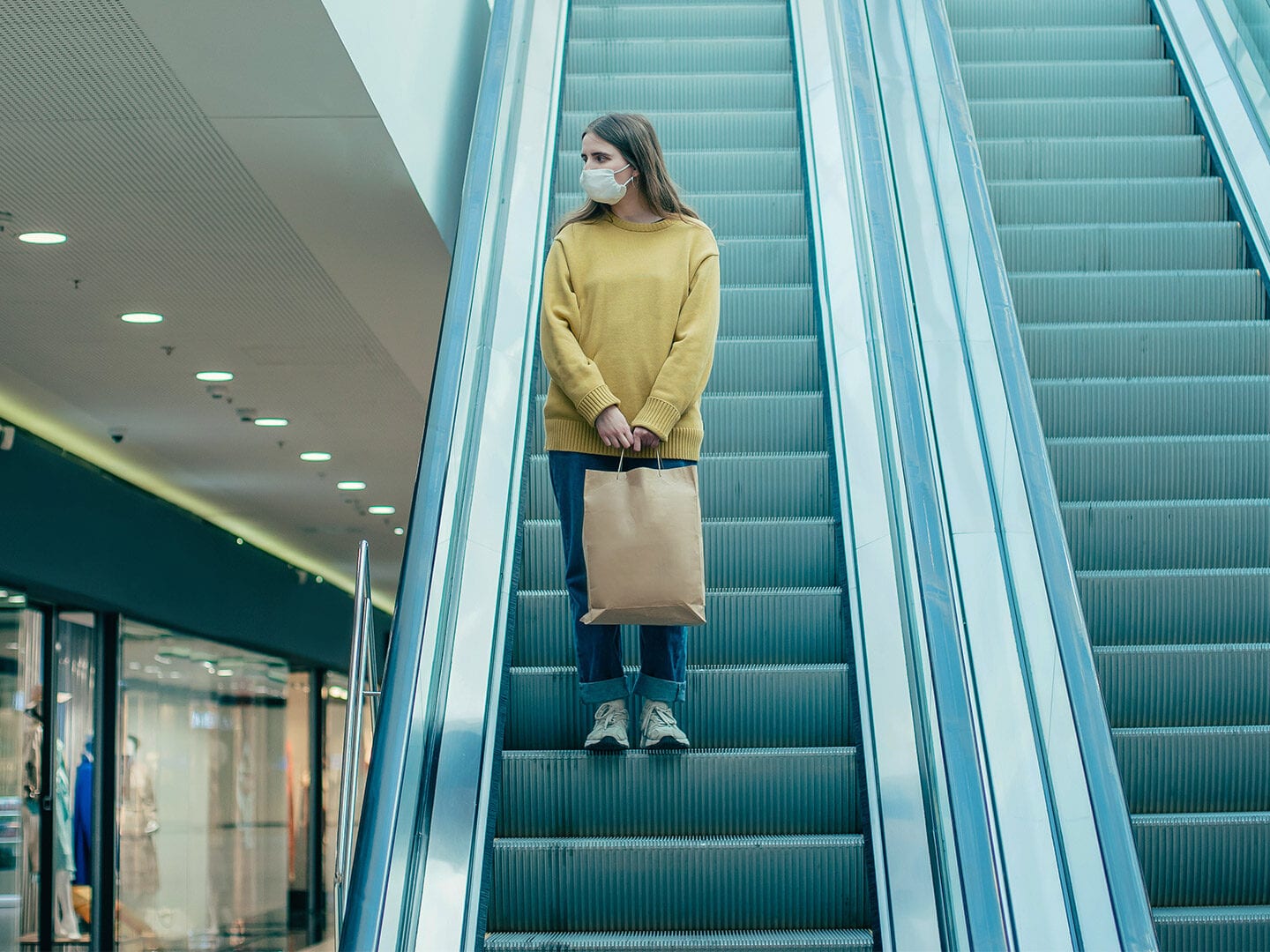 EIne Frau steht alleine auf einer Rolltreppe mit einer Atem-Schutz-Maske im Gesicht