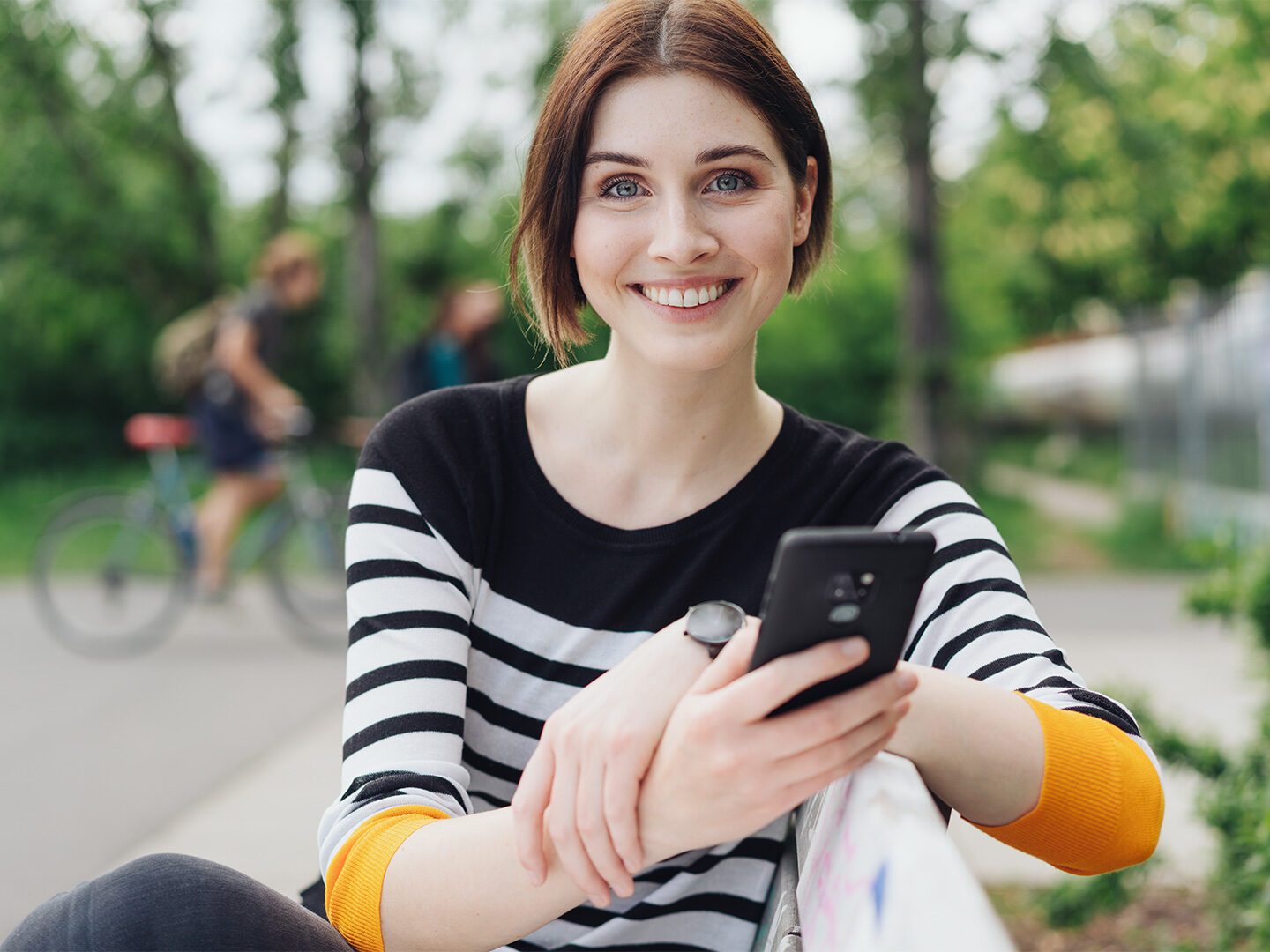 Junge Frau mit Smartphone in der Hand sitzt auf einer Bank auf einem Schulhof. Im Hintergrund sind Fahrradfahrer zu sehen.