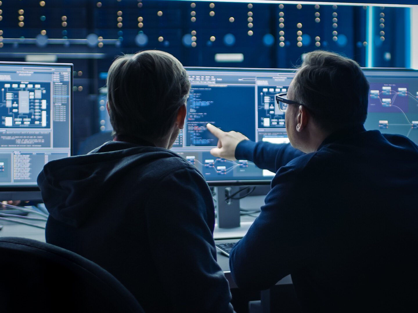 Titelbild IT-Sicherheit: Zwei Männer sitzen in einem Raum mit mehreren Computerbildschirmen und schauen sich einen Code an