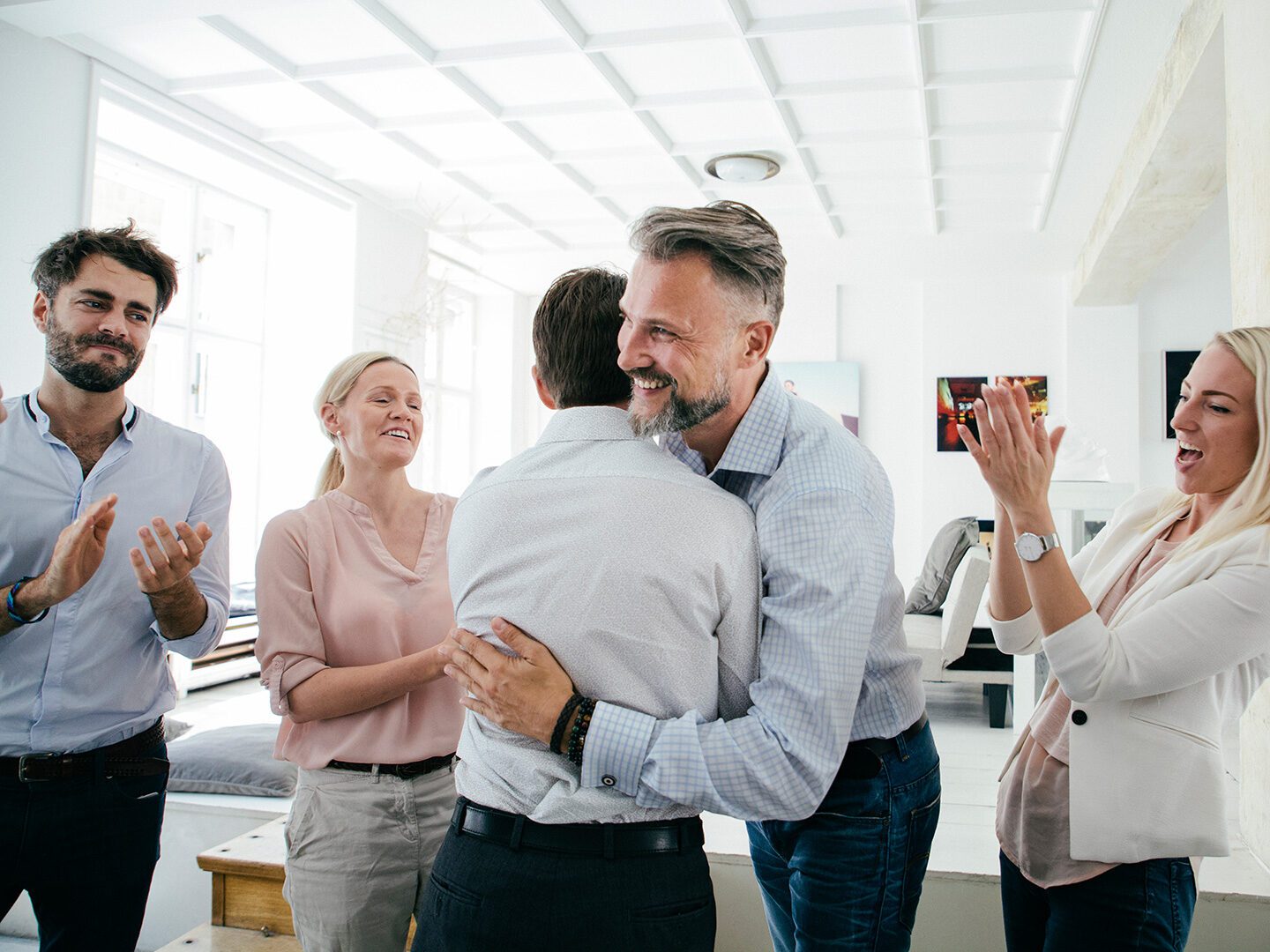 Bürosituation, in der sich zwei Männer fröhlich umarmen und die Kolleginnen drumherum applaudieren.
