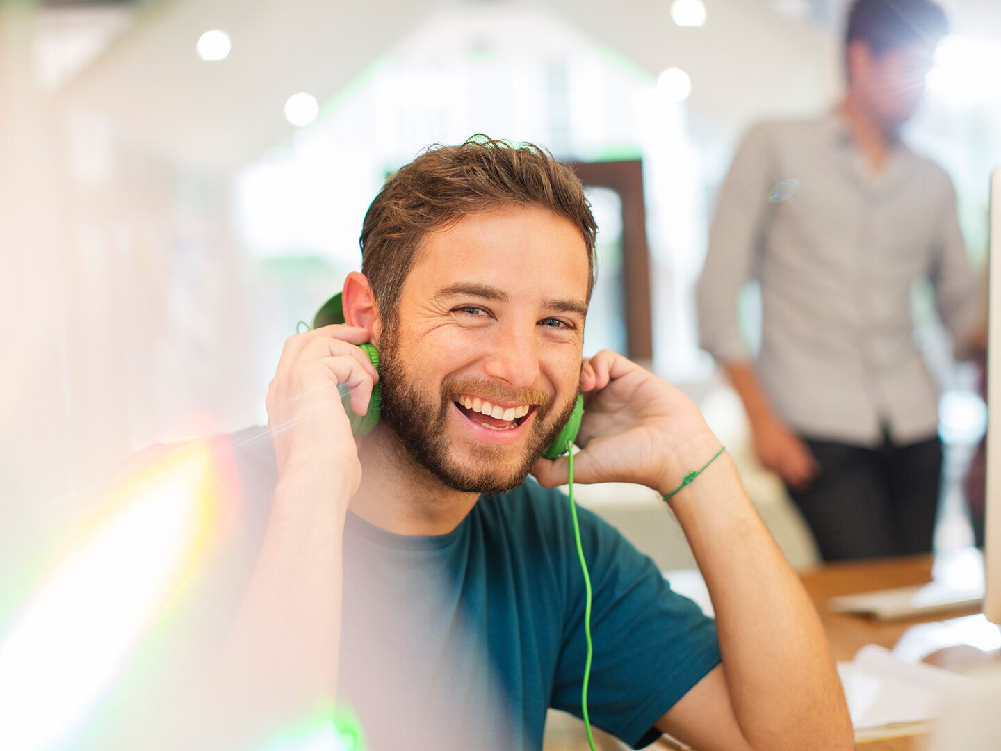 ein junger Mann sitze in einer Büro-Umgebung, nimmt seine grünen Kopfhörer ab und lächelt in die Kamera