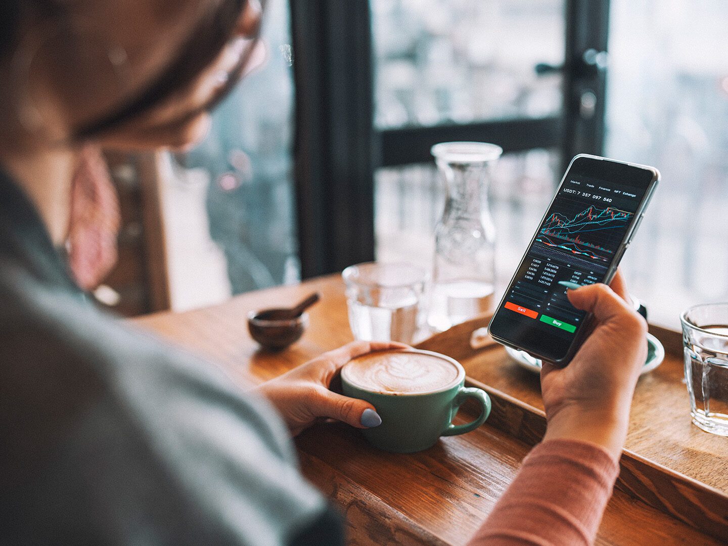 eine Frau trinkt Kaffee in einem Café und checkt dabei den Aktienkurs auf dem Smartphone